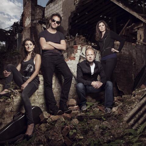 Four band members in dark clothing posed amongst the ruins of a brick building