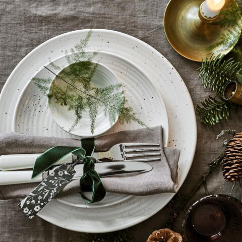 Christmas table setting with white crockery, a rolled white linen napkin and green foliage 